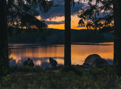 Kompisgäng vid lägereld intill sjö i solnedgång