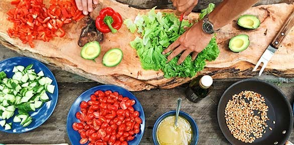 Träbord med stor skärbräda av trä med sallad, paprika och avokado och skålar med gurka, tomat och pinjenötter