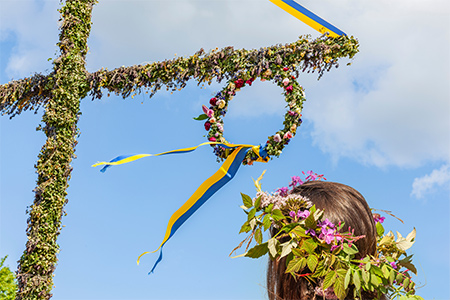 Kvinna med blomsterkrans på huvudet blickar upp mot en midsommarstång