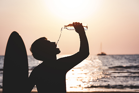 Kille med surfbräda dricker vatten ur vattenflaska i solnedgång