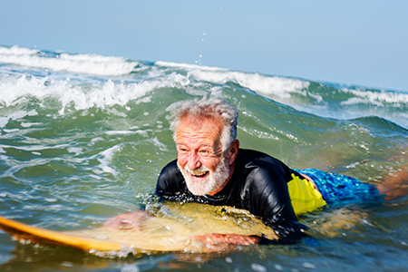 Äldre man med vitt hår och skägg paddlar på surfbräda i havet