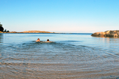 Två personer simmar ut i havet från en sandstrand. Längre ut i vattnet skymtar klippor.