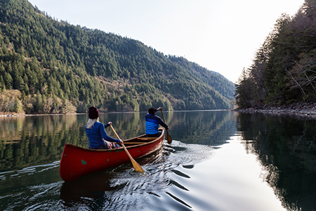 Två personer paddlar kanot i ett stilla vatten, på sidorna sluttar skogen ner mot vattnet. 