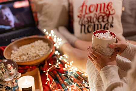 Två händer håller en kopp varm choklad med marshmallows på toppen. I bakgrunden en datorskärm med julfilm.