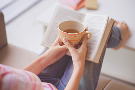 En kvinna sitter i en soffa och läser bok med en tekopp i handen