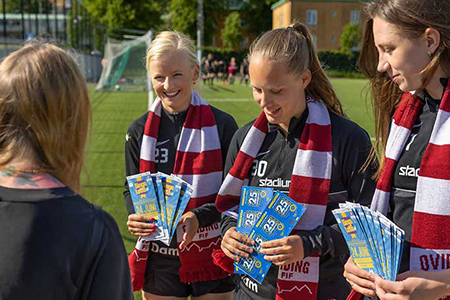 Tre kvinnor i fotbollsställ säljer bingolotter och sverigelotter ute i solen