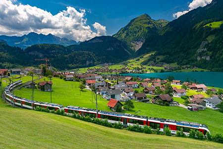 Ett rött tåg kör genom ett somrigt alplandskap med en by i bakgrunden